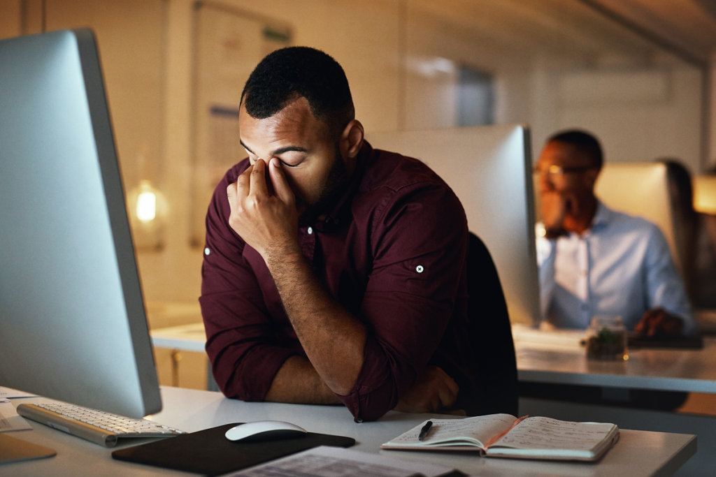 Homem em sua mesa de trabalho leva uma mão ao rosto cansado. Sinais de desgaste no trabalho.